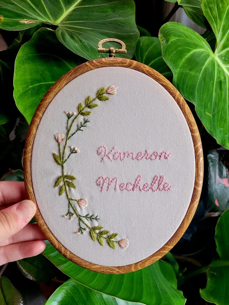 Hand-embroidered hoop with a floral sprig and the name Kameron Michelle, held against houseplant leaves